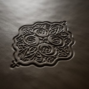 May include: A close-up of a brown leather surface with an intricate embossed design in the center. The design features a repeating pattern of circles and swirls.