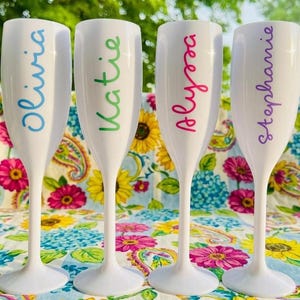 May include: Four white plastic champagne flutes standing in a row. Each flute features a name printed vertically in a different colour: Olivia in blue, Katie in green, Alyssa in pink, and Stephanie in purple. The flutes have a smooth finish.
