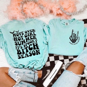 Puede incluir: Dos camisetas azul claro con gráficos negros. Una camiseta dice "Move Over Hot Girl Summer It's Spooky Bitch Season". La otra camiseta tiene una mano de esqueleto con el texto "Spooky Bitch Seasons".