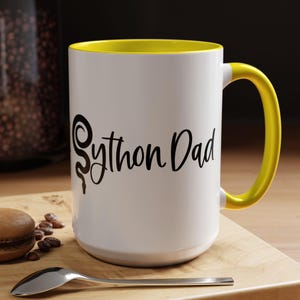 Op de afbeelding: Een witte keramische mok met een gele rand en handvat. De mok heeft een zwart slangontwerp en de tekst "Python Dad" erop gedrukt.