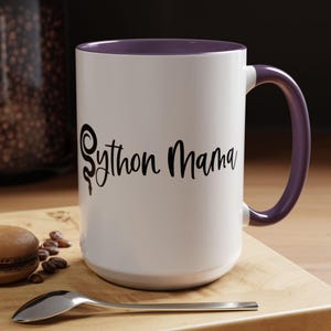Könnte beinhalten: Weißer Keramikbecher mit lila Innenraum und Henkel. Der Becher trägt den Text "Python Mama" in schwarzer Schrift. Der Becher steht auf einer Holzoberfläche mit einem Löffel, Kaffeebohnen und Gebäck.