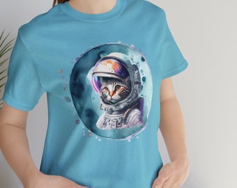 May include: A light blue t-shirt with a watercolor illustration of a cat wearing an astronaut helmet. The cat is looking to the side and has a serious expression. The illustration is surrounded by a circle of white dots.