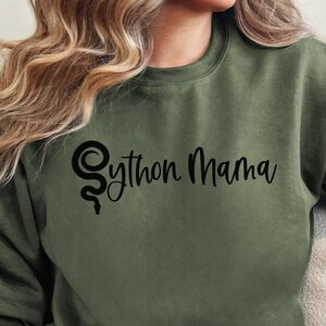 Può includere: Una felpa verde con la scritta "Python Mama" e un design di serpente stilizzato in nero.