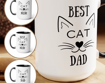 Puede incluir: Taza de café de cerámica blanca con borde y asa negros. La taza tiene una cara de gato negra con una nariz en forma de corazón y el texto "BEST CAT DAD".
