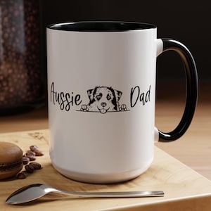 May include: White ceramic mug with a black rim and handle. The mug features a black and white illustration of an Australian Shepherd dog peeking over the top with the text "Aussie Dad" printed on the mug.