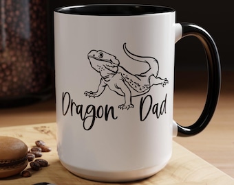May include: White ceramic mug with a black rim and handle. The mug has a black and white illustration of a bearded dragon and the text "Dragon Dad".