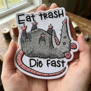 May include: Embroidered patch featuring a gray opossum with pink accents, lying on its back. The patch has the text "Eat trash Die Fast" in black lettering on a white background. The patch is held in a person's hands.