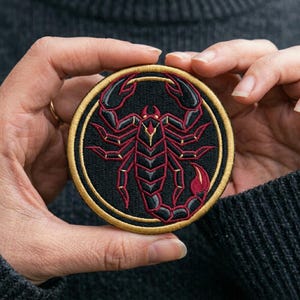 May include: Embroidered patch featuring a scorpion design. The patch has a black background with a red and gold scorpion. The scorpion is surrounded by a gold border. The patch is round.