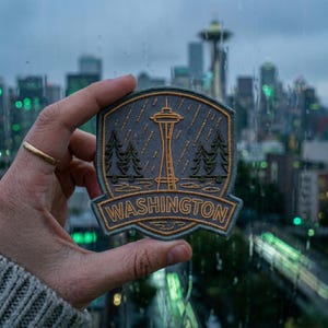 May include: A gray and gold embroidered patch featuring the Seattle Space Needle, pine trees, and rain. The word "WASHINGTON" is embroidered in gold at the bottom of the patch. The patch is held in front of a blurred city skyline.