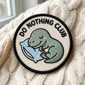 May include: Embroidered patch with a black border and the text "DO NOTHING CLUB". The patch features a green dinosaur sleeping on a blue pillow with white stars. The patch is on a cream-colored knitted surface.