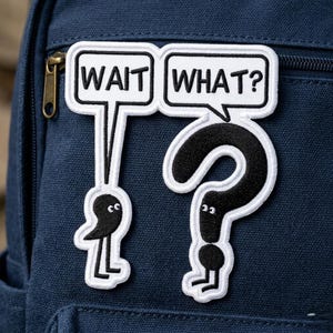 May include: Embroidered patch with the words "WAIT WHAT?" in a speech bubble design. The patch features a black question mark and two cartoon figures with black eyes and white outlines, attached to a navy blue backpack.