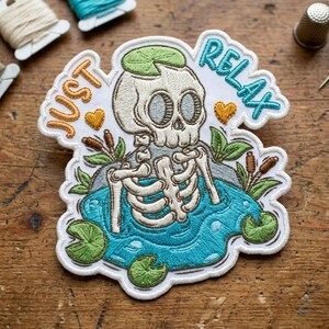 May include: Embroidered patch featuring a skeleton in a pond with lily pads and cattails. The patch has the words "JUST RELAX" in orange and blue. The skeleton has a green lily pad on its head and two orange hearts.