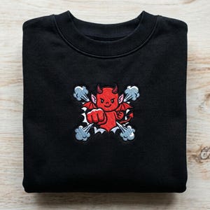 May include: Black sweatshirt featuring a red devil graphic with horns, wings, and a clenched fist. The devil is surrounded by stylized gray smoke. The sweatshirt is folded neatly on a light wood surface.