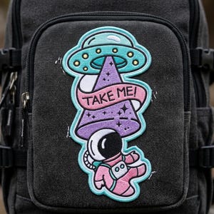 May include: Embroidered patch featuring an astronaut being beamed up by a UFO. The design includes a purple beam, a teal UFO, and the text "TAKE ME!" in a banner. The astronaut is wearing a pink spacesuit.