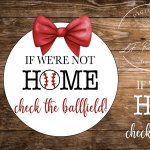 May include: A circular white sign featuring a large red bow at the top and black text that reads IF WE'RE NOT HOME check the ballfield! with a baseball graphic replacing the letter O in HOME. The design is presented on a dark wood background.