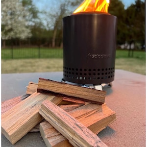 May include: A cylindrical black Solo Stove fire pit with a row of ventilation holes near the base, emitting bright orange flames from the top. A small pile of rectangular, light brown wooden kindling sticks is arranged in the foreground.