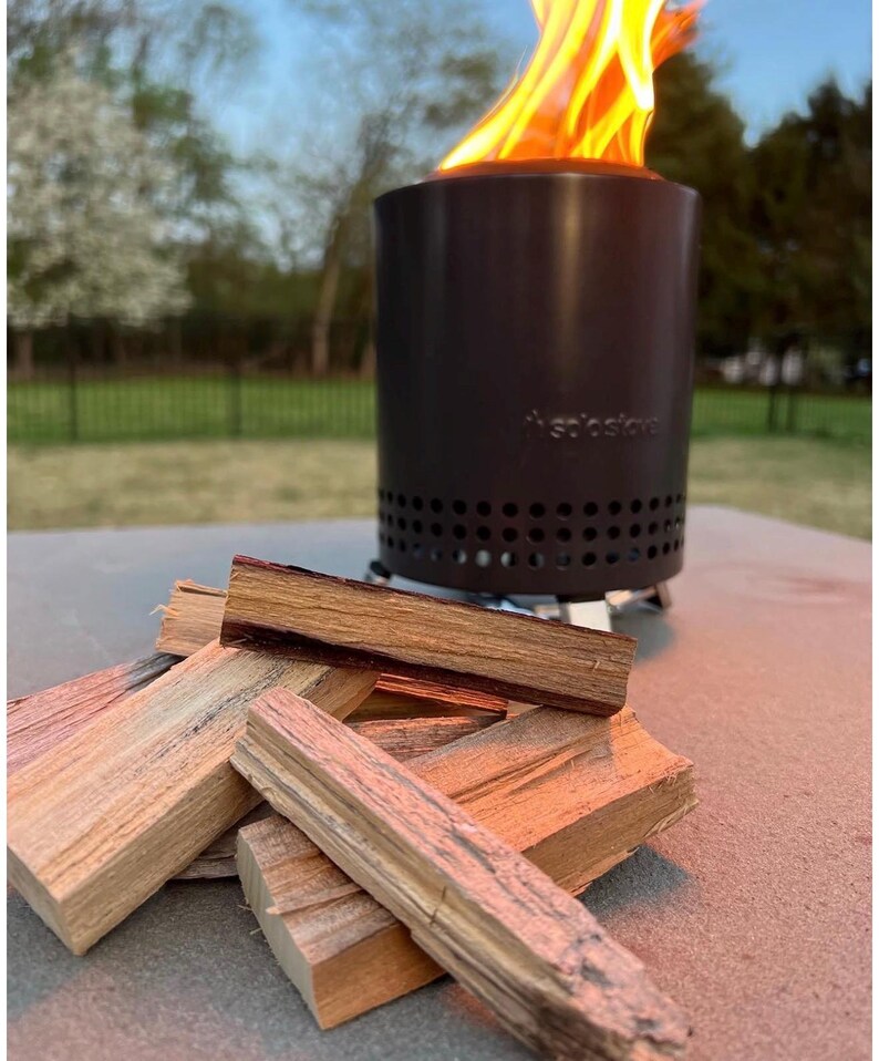 May include: A cylindrical black Solo Stove fire pit with a row of ventilation holes near the base, emitting bright orange flames from the top. A small pile of rectangular, light brown wooden kindling sticks is arranged in the foreground.