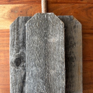 May include: A pumpkin-shaped decorative object constructed from three vertical pieces of weathered gray wood. The central piece is taller and features a small, cylindrical wooden stem attached to the top edge, showcasing a rough, textured grain.