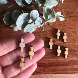 May include: Small, wooden figurines in the shape of a person with a crown. The figurines are light brown with a darker brown outline. The figurines are on a wooden surface with green leaves in the background.