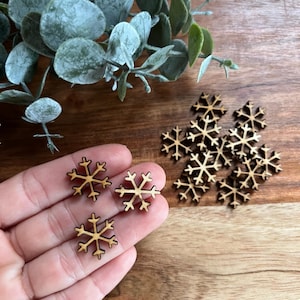 May include: Wooden snowflake cutouts in a natural wood tone. Three snowflakes are held in a hand, while a small pile of snowflakes sits nearby. Greenery is visible in the background. These decorative snowflakes are perfect for crafts.