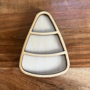 May include: A wooden candy corn shaped tray with three sections. The tray is made of light-colored wood with a dark outline. The tray is sitting on a wooden surface.