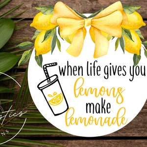 May include: A white circle with a yellow bow and lemons around the edge. The text "when life gives you lemons make lemonade" is written in black. A black and white illustration of a glass of lemonade with a straw is in the lower left corner.