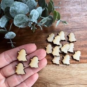 May include: A collection of small, wooden ghost-shaped cutouts with dark brown outlines. Some are held in a hand, while others are scattered on a wooden surface. Greenery is visible in the background.
