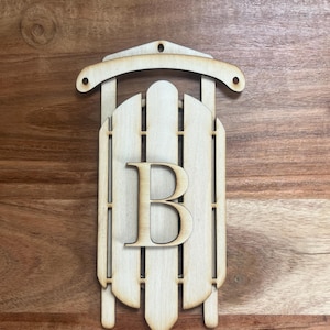 May include: A wooden sled-shaped ornament with the letter "B" in the center. The ornament is made of light-colored wood and has a decorative design. It features a curved top with a hole for hanging.