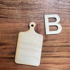 May include: A small, unfinished wooden cutting board with a handle and a hole for hanging, next to a wooden letter "B". The items are light-colored and sit on a brown wooden surface.