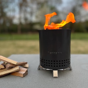 May include: A black cylindrical Solo Stove portable fire pit with a row of circular ventilation holes near the base and silver metal legs. Bright orange flames rise from the top of the unit, and a small pile of light brown wood kindling sits nearby.