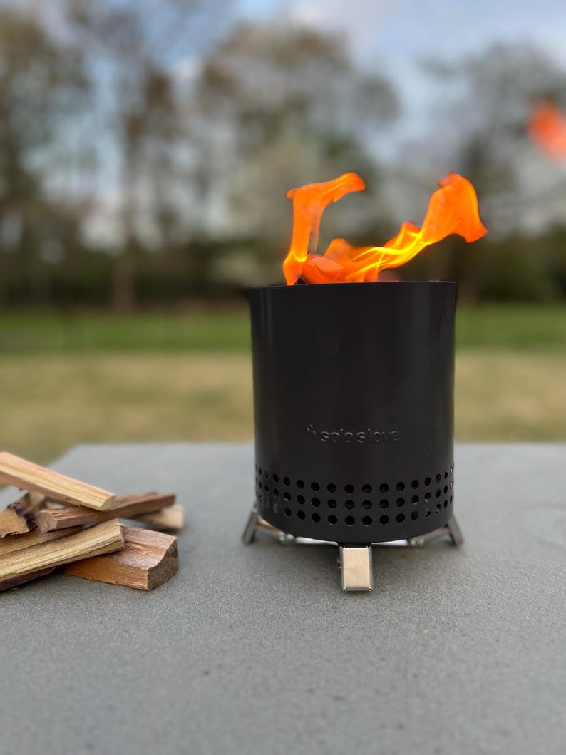 May include: A black cylindrical Solo Stove portable fire pit with a row of circular ventilation holes near the base and silver metal legs. Bright orange flames rise from the top of the unit, and a small pile of light brown wood kindling sits nearby.