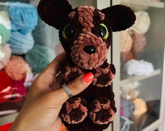 May include: A handmade brown and tan crocheted dog plushie with large green eyes and a black nose. The plushie has a textured, velvety appearance and is held in front of a shelf filled with colorful yarn.