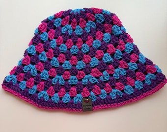 May include: A handmade crochet bucket hat in a granny square pattern. The hat features a color scheme of purple, hot pink, and turquoise blue. The brim is a solid dark pink. A small tag with text is attached to the hat.