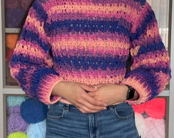 May include: A colorful, handmade crochet sweater in shades of pink, purple, and blue. The sweater features a boat neck and long sleeves. The yarn appears to be soft and textured, perfect for a cozy, handmade look. The sweater is a unique piece of wearable art.