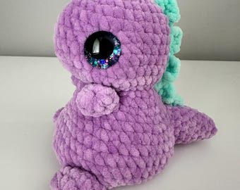 May include: A handmade, plush purple dinosaur with large, glittery eyes. The crochet dinosaur has mint green accents on its back and head. The soft toy is designed in a sitting position, showcasing its small arms and tail. A cute, cuddly, and unique gift.