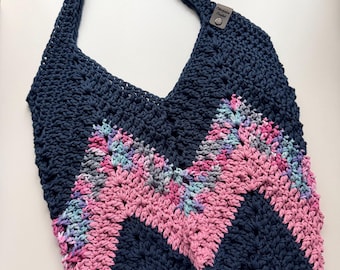 May include: A navy blue crochet tote bag with a chevron design in pink, gray, and blue. The bag has a single strap and a small tag near the top. The handmade bag is suitable for carrying items.
