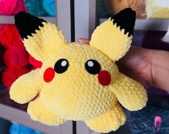 May include: A yellow crocheted Pikachu plush toy with black ears and eyes, and red cheeks. The toy is held in a hand against a background of colorful yarn balls.
