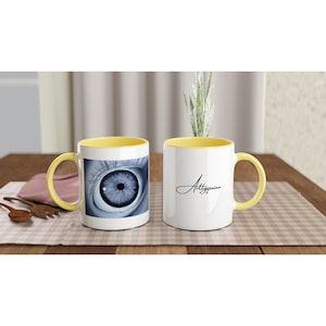 May include: Two white ceramic mugs with yellow interiors and handles. One mug features a black and white close-up graphic of a human eye. The other mug displays the black cursive text Arttyquinn on the front. Both mugs have a cylindrical shape.