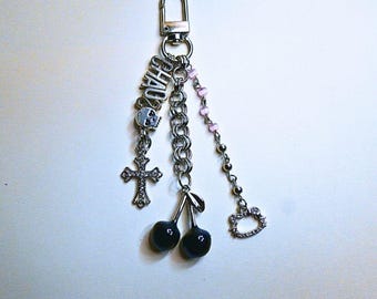 May include: Silver-toned keychain with multiple charms. Charms include a cross, a skull, the word "CHAOS", cherries, and a Hello Kitty-shaped charm. Pink beads and a chain are also attached. The keychain is attached to a silver clasp.