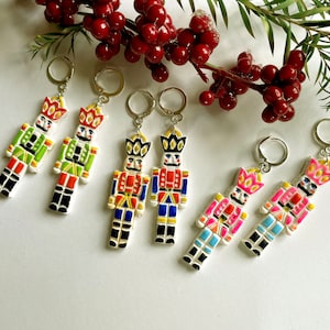 May include: Six pairs of colourful nutcracker earrings. Each pair features a different design with a white background and colourful details. The earrings are made of polymer clay and are attached to silver ear wires.