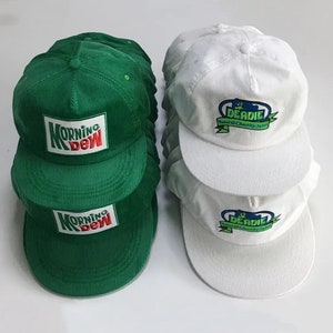 Puede incluir: Una pila de gorras de béisbol de pana verde con el texto "Morning Dew" en rojo y blanco. Una pila de gorras de béisbol de pana blanca con el texto "Deaddie Natural Chunky Jams" en azul, verde y blanco.