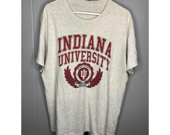 Könnte beinhalten: Hellgraues T-Shirt mit dem Schriftzug "INDIANA UNIVERSITY" in roten Buchstaben. Darunter befindet sich ein weinrotes Emblem mit einem "U"-Symbol und dem Wort "HOOSIERS". Das T-Shirt hängt an einem weißen Kleiderbügel.