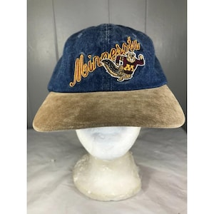 May include: A blue denim baseball cap featuring the word Minnesota embroidered in yellow cursive script and a mascot graphic of a gopher wearing a maroon and gold jersey. The cap has a solid tan suede brim and a structured crown design.