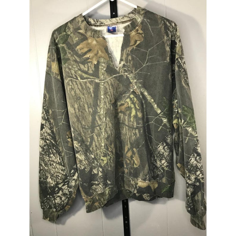 May include: A long-sleeved camouflage sweatshirt with a V-neck. The pattern features shades of green, brown, and tan, mimicking a forest environment. The sweatshirt is hanging on a white hanger against a white background.