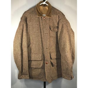 Puede incluir: Chaqueta de tweed marrón con cuello de pana y múltiples bolsillos. La chaqueta tiene cierre de botones y está colgada de una percha blanca. La chaqueta parece ser de estilo vintage.