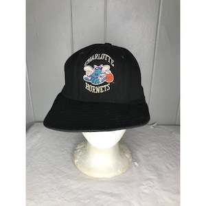 Puede incluir: Gorra de béisbol negra con un logotipo de los Charlotte Hornets que presenta un avispón de dibujos animados sosteniendo una pelota de baloncesto. El logotipo está bordado en amarillo, verde, azul y naranja.