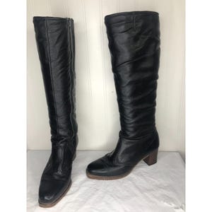 May include: A pair of black knee-high boots. One boot has a zipper on the side. Both boots have a rounded toe, a wooden heel, and a rubber sole. The boots are made of a smooth, shiny material.