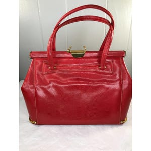 Puede incluir: Un bolso de mano vintage rojo con un patrón texturizado similar al de un reptil. El bolso presenta un cierre y detalles dorados, con dos asas superiores. El bolso, de forma rectangular, tiene un diseño estructurado y probablemente está hecho de cuero o material similar.