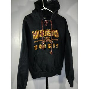May include: Black hooded sweatshirt with the words "MINNESOTA HOCKEY" in gold and maroon lettering. The sweatshirt has a front pocket and a drawstring with maroon and gold accents. The hood is up, and the sweatshirt is hanging on a white hanger.