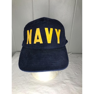 May include: Navy blue corduroy cap with the word "NAVY" in yellow block letters across the front. The cap has a curved brim and a structured crown. The hat is likely a vintage or military-inspired accessory.